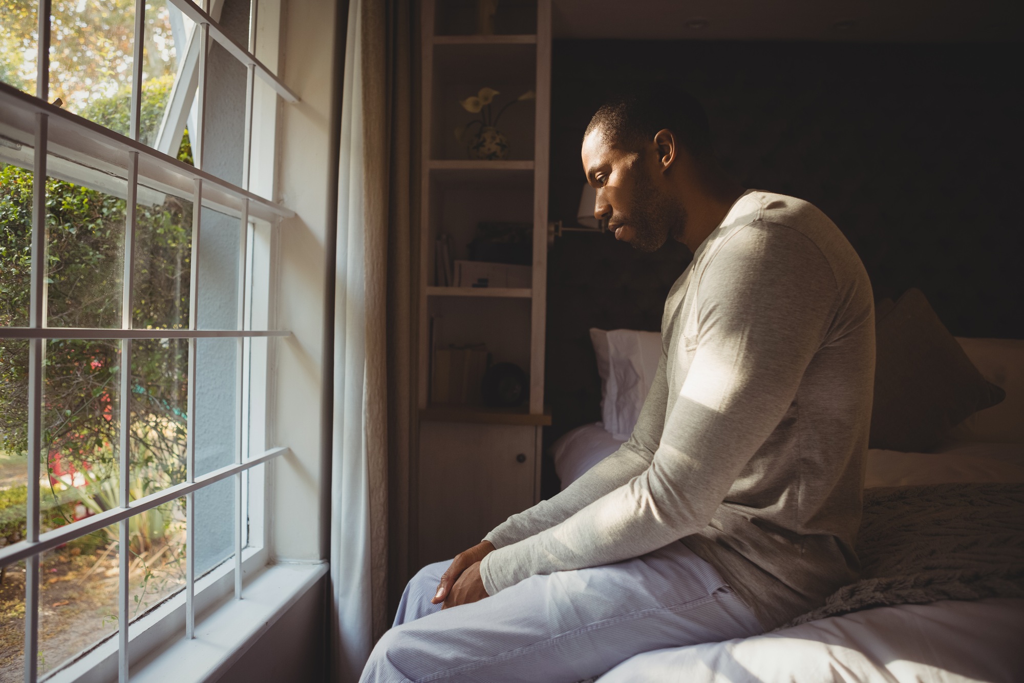 Side view of sad man sitting on bed by window